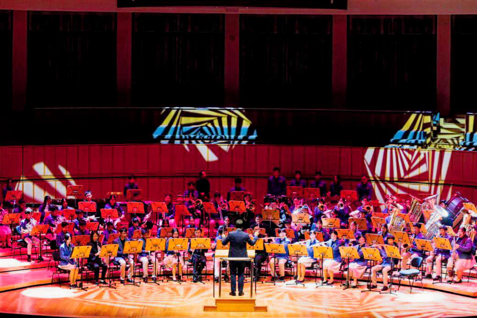 Photograph of the BDS Symphonic Band performing on stage in a concert hall with a conductor leading from the center. Stage lighting includes patterned spotlights in yellow and white, highlighting musicians seated in rows with various instruments and music stands. 